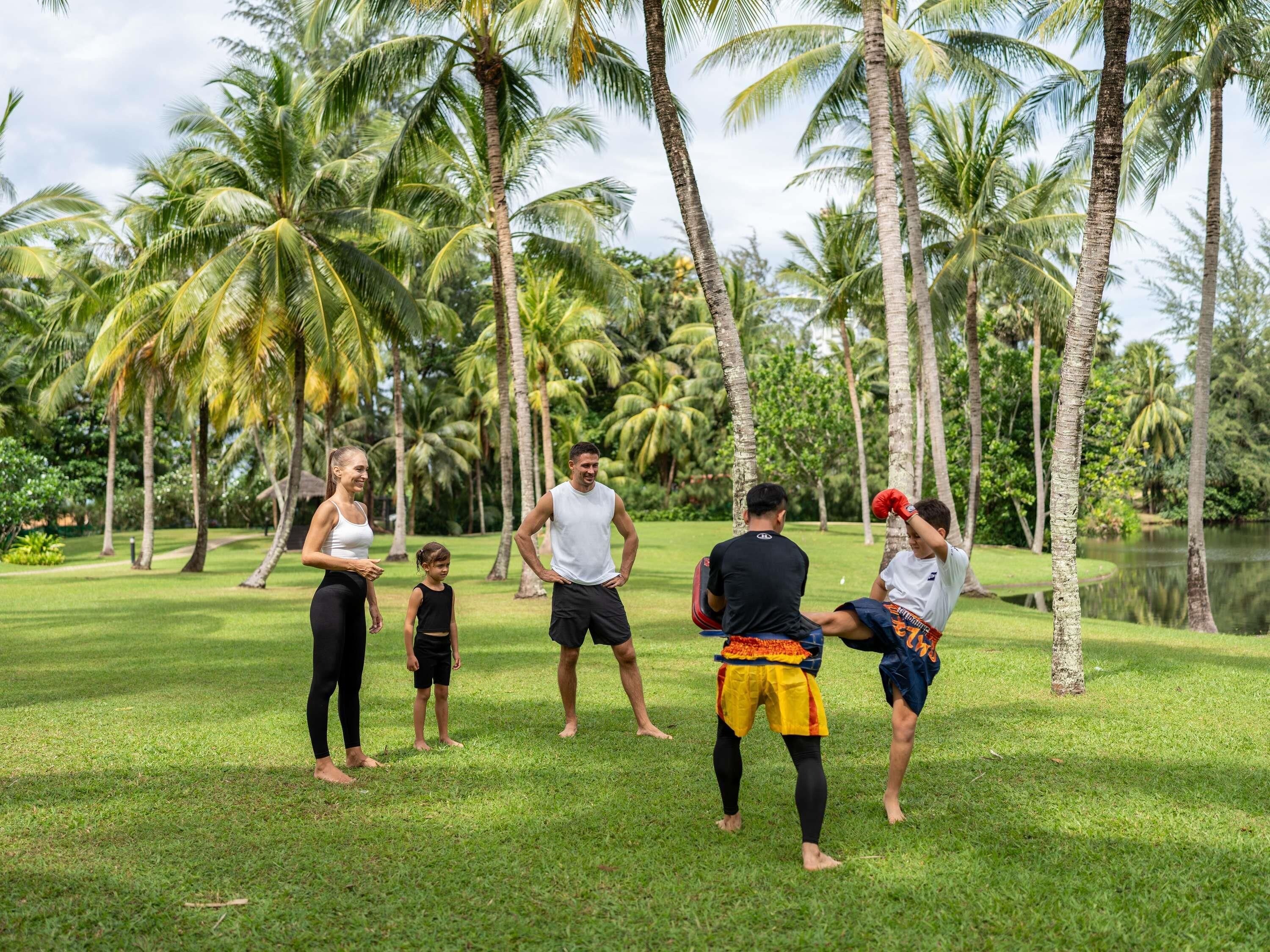 Un grupo de personas en un área verde al aire libre, con palmeras y un cuerpo de agua en el fondo.