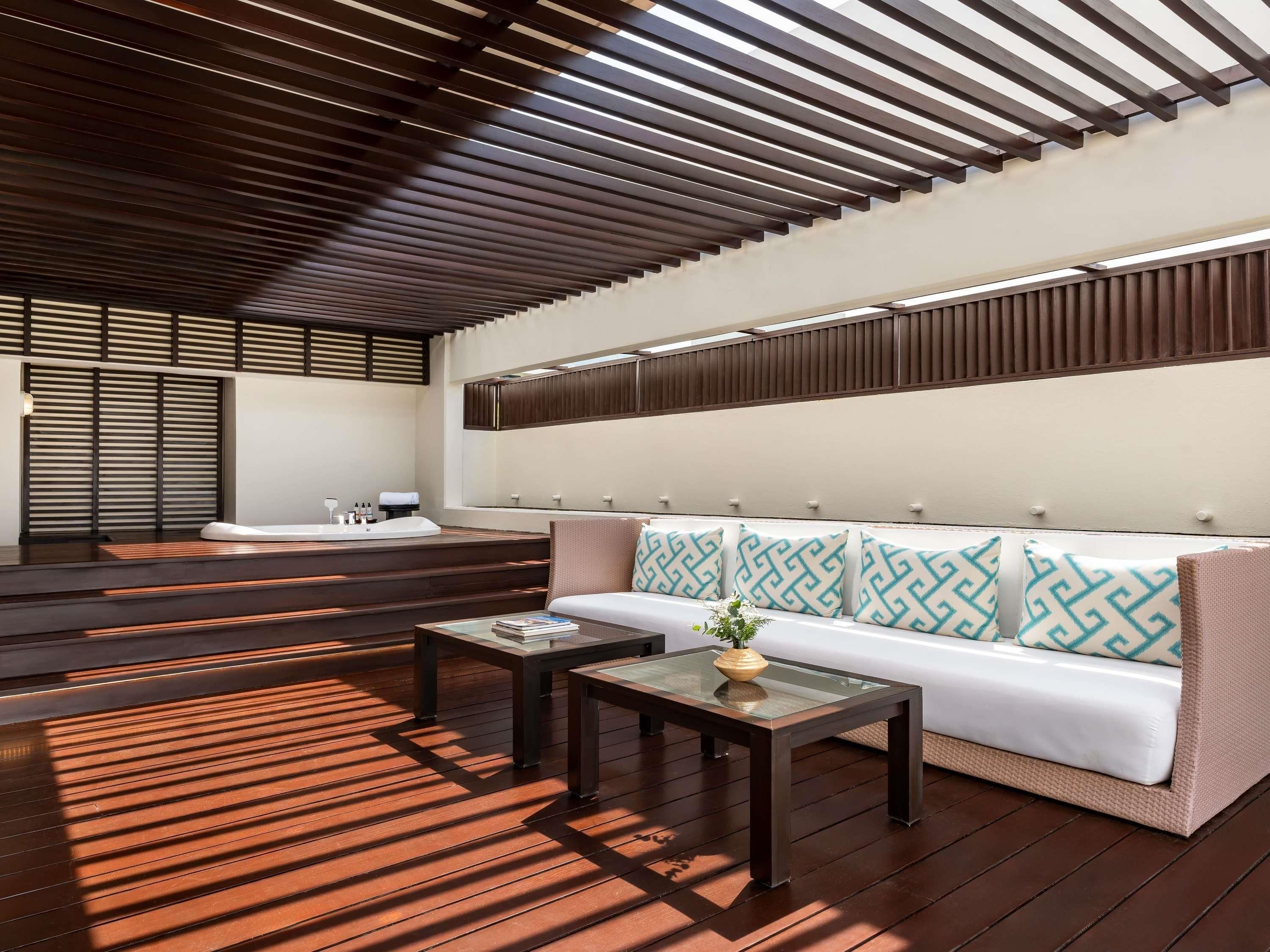 A modern bathroom with a wooden floor, a bathtub, a sofa with patterned cushions, and a glass-top table with a vase.