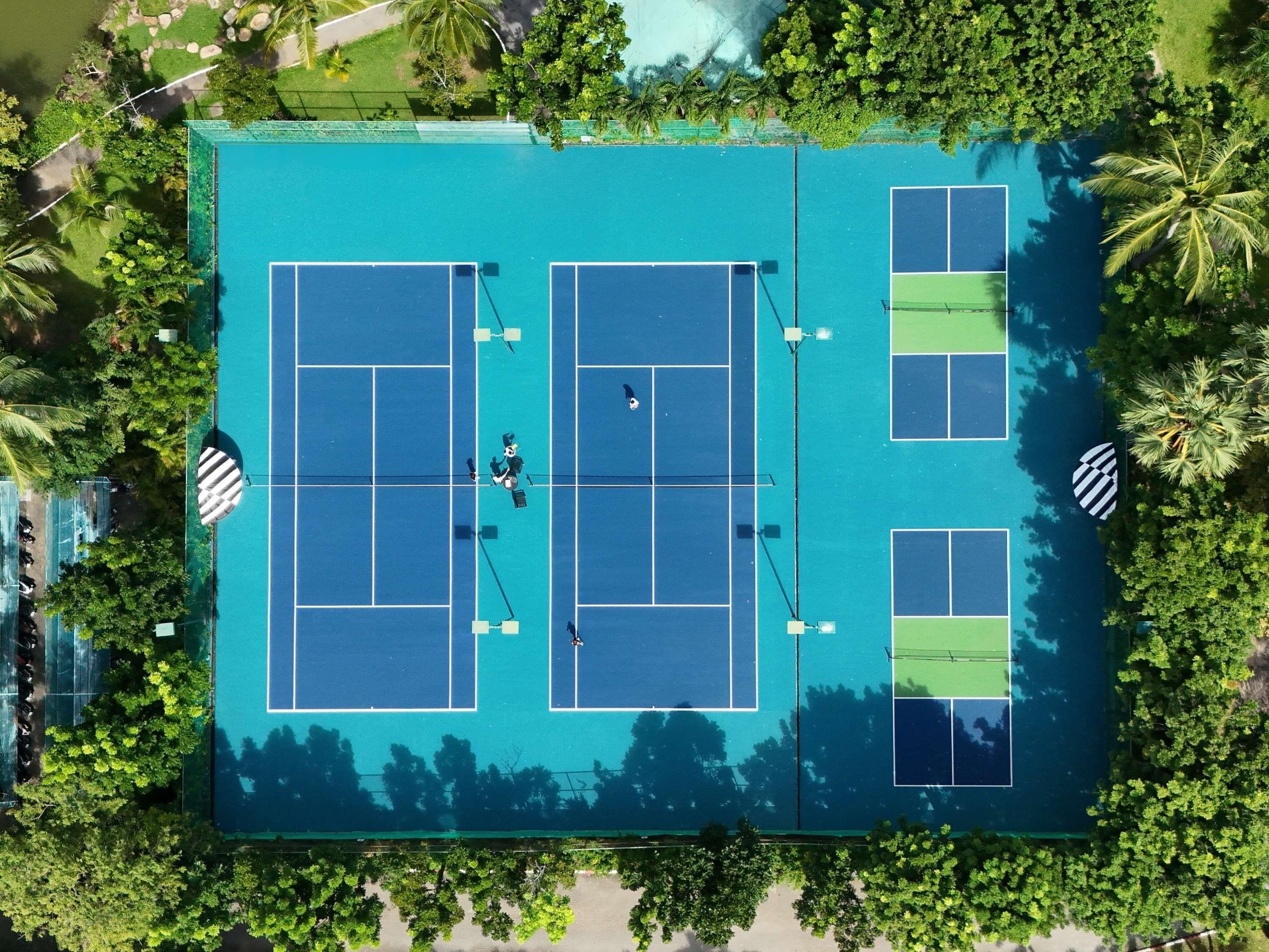 An aerial view of a tennis court complex with multiple courts, surrounded by lush greenery and palm trees.