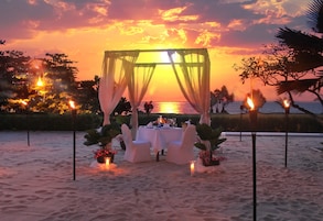 A beachside dining setup with a canopy, white chairs, and tables, overlooking the sea.