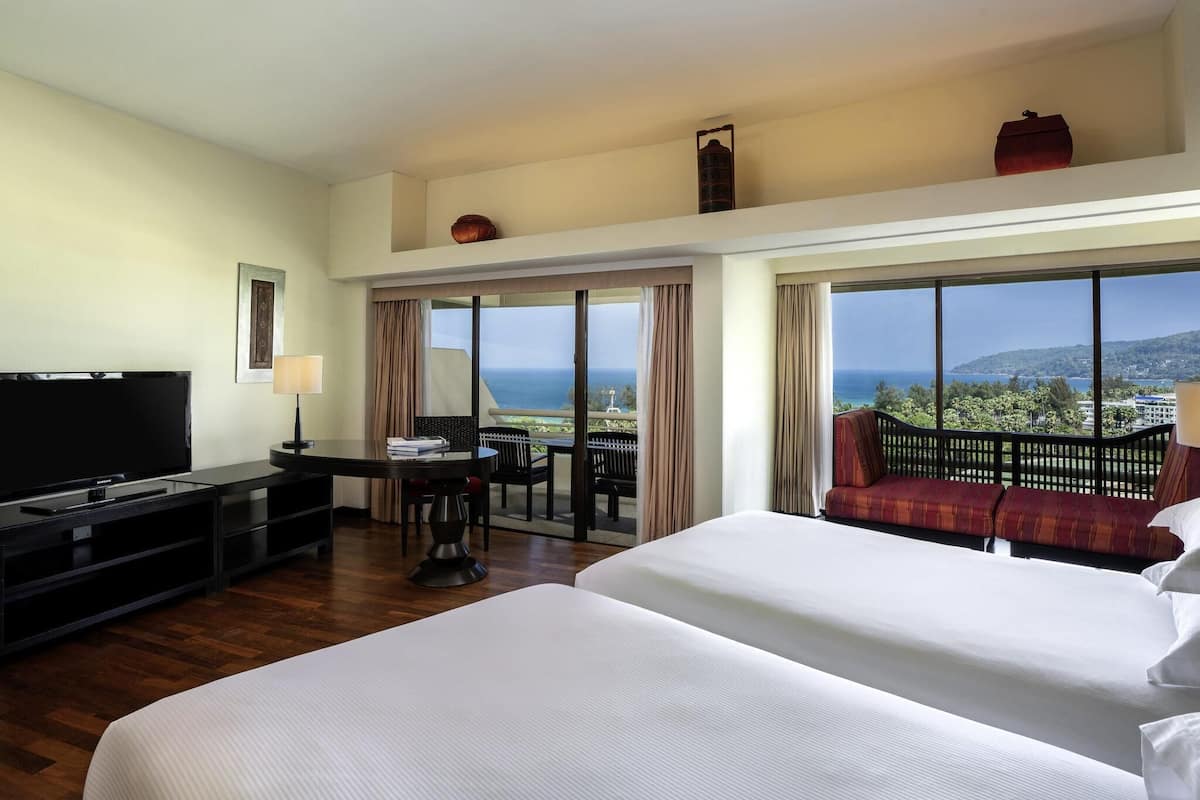 Une chambre dâhĂŽtel Ă©quipĂ©e dâune tĂ©lĂ©vision, dâun bureau avec une chaise, de deux lits, dâun balcon avec vue sur la mer et dâune grande fenĂȘtre.