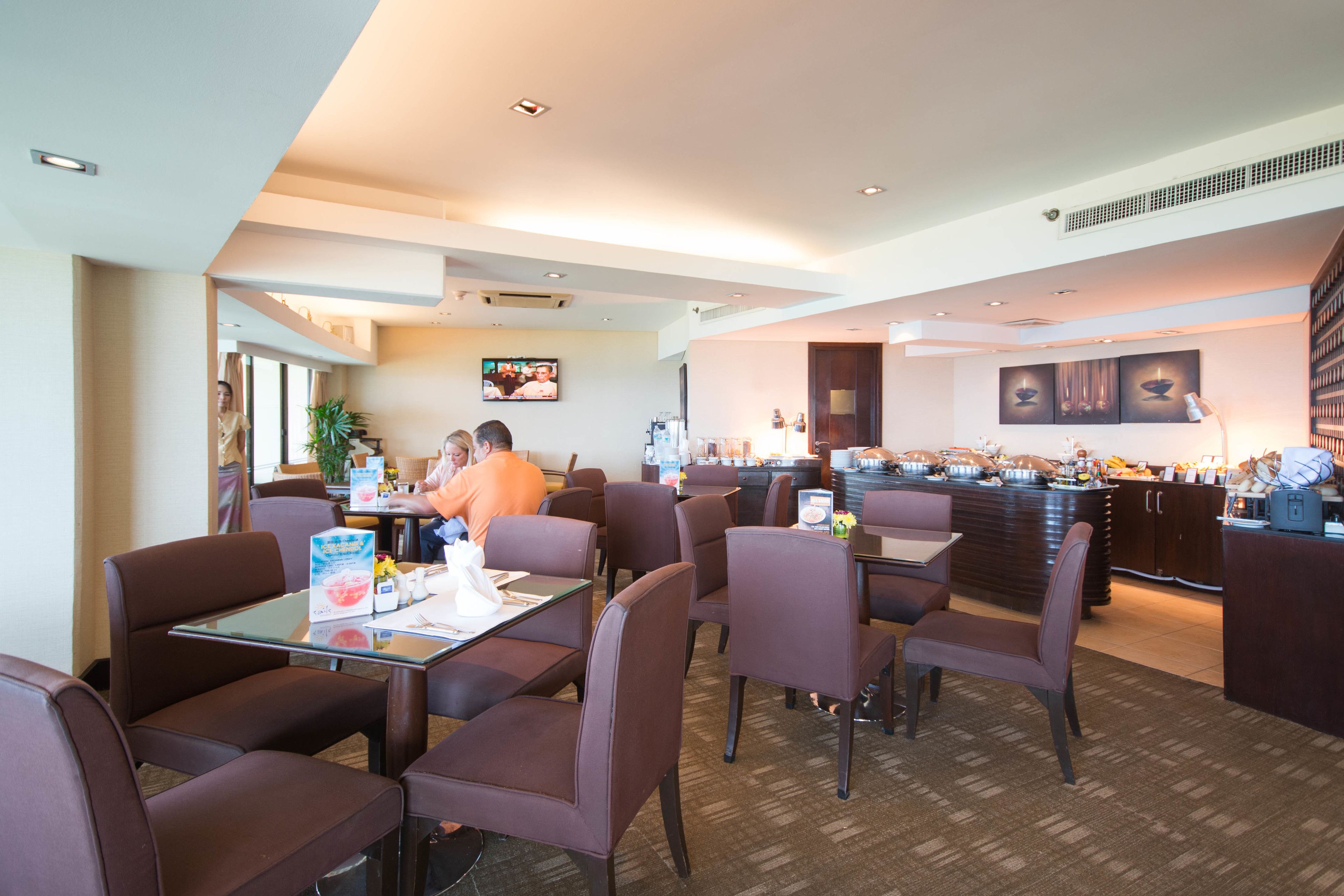 A hotel dining area with multiple tables and chairs, a buffet counter, and a television screen.