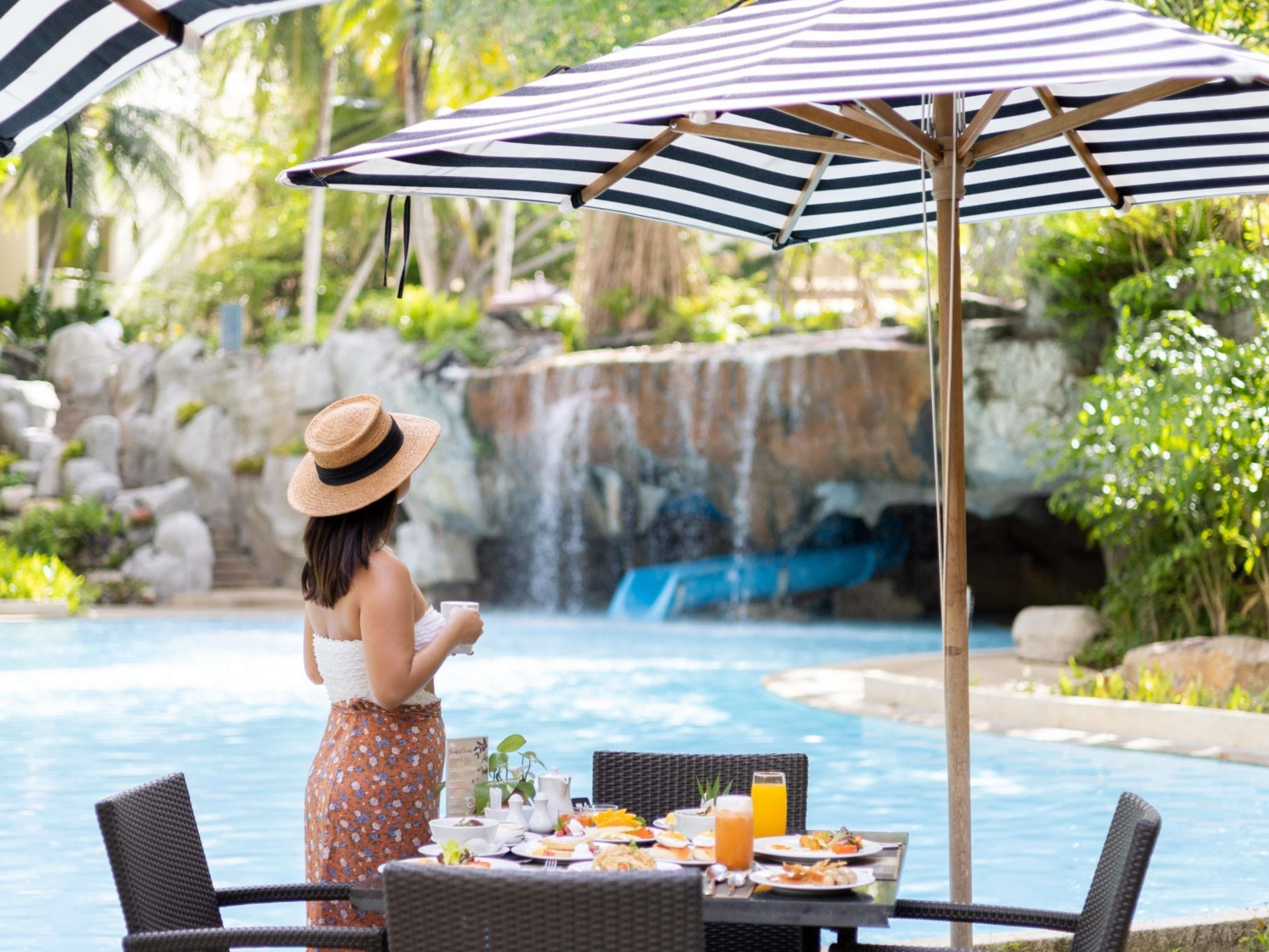Une personne est assise à une table au bord de la piscine, devant un repas, et a une vue sur une cascade.