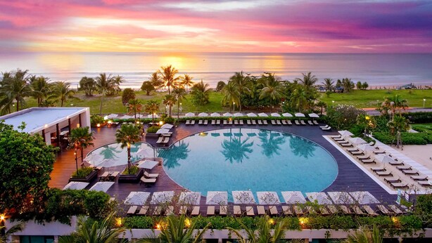 A resort pool with lounge chairs and umbrellas, surrounded by palm trees and a view of the ocean at sunset. - Pullman Phuket Karon Beach Resort (Karon)