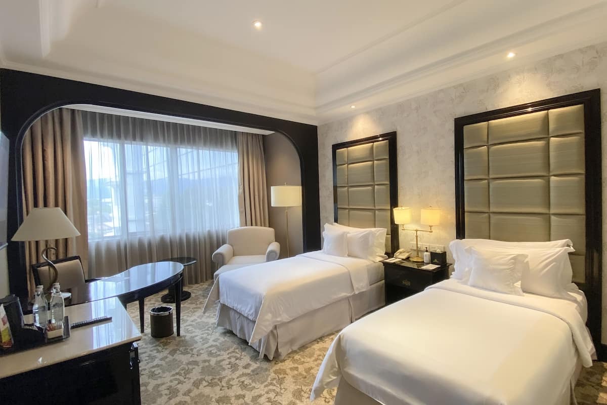 A hotel room with two beds, a desk, a chair, a lamp, and a nightstand with a phone and lamps.