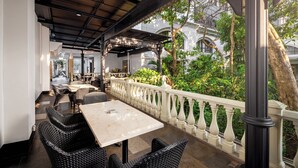 Un área de estar al aire libre con muebles de mimbre y una mesa blanca, con vistas a un jardín exuberante y un edificio con ventanas arqueadas.