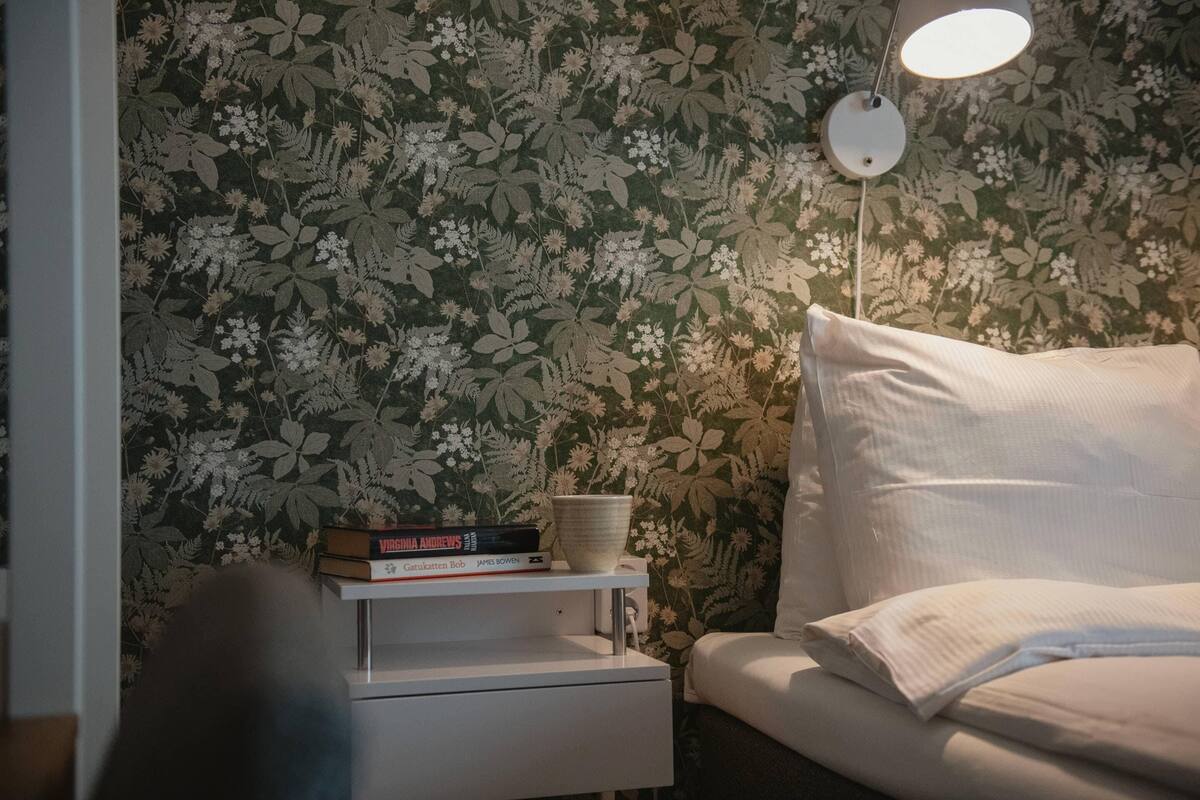 A bedroom with floral wallpaper, a bedside table with books and a bowl, a white bed with a pillow, and a hanging lamp.