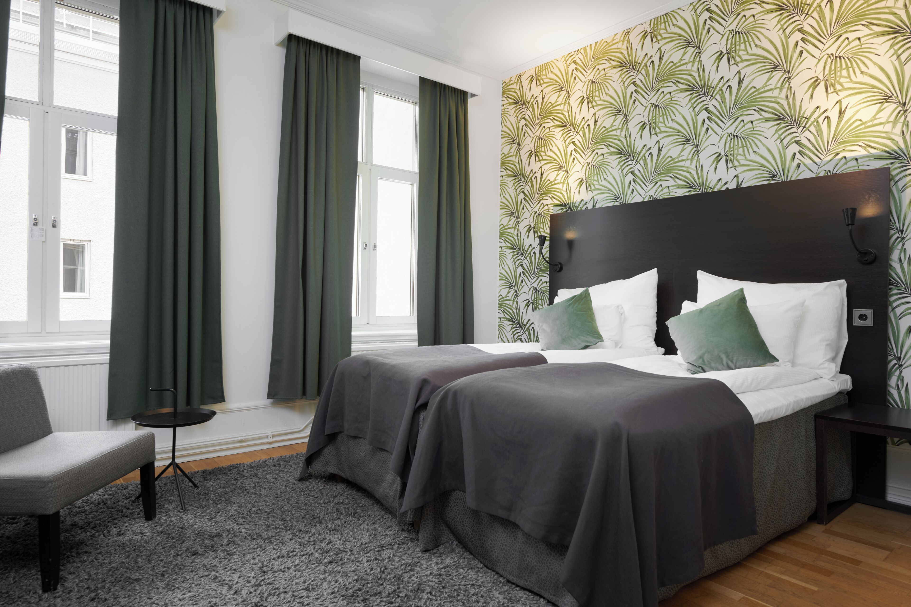 Habitación de hotel con dos camas, una silla, una mesita pequeña y un papel tapiz estampado.