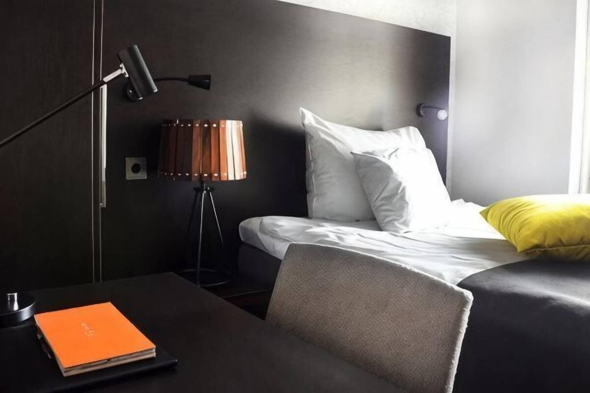 A hotel room with a bed, a desk with a lamp, a chair, and a small table with a book.