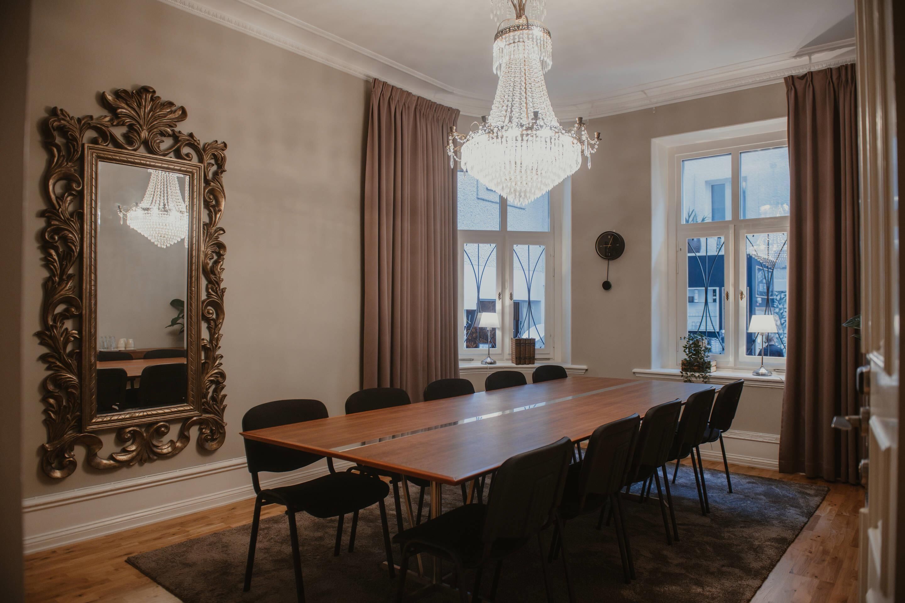A dining room with a wooden table, black chairs, a large ornate mirror, a chandelier, and a view through large windows.