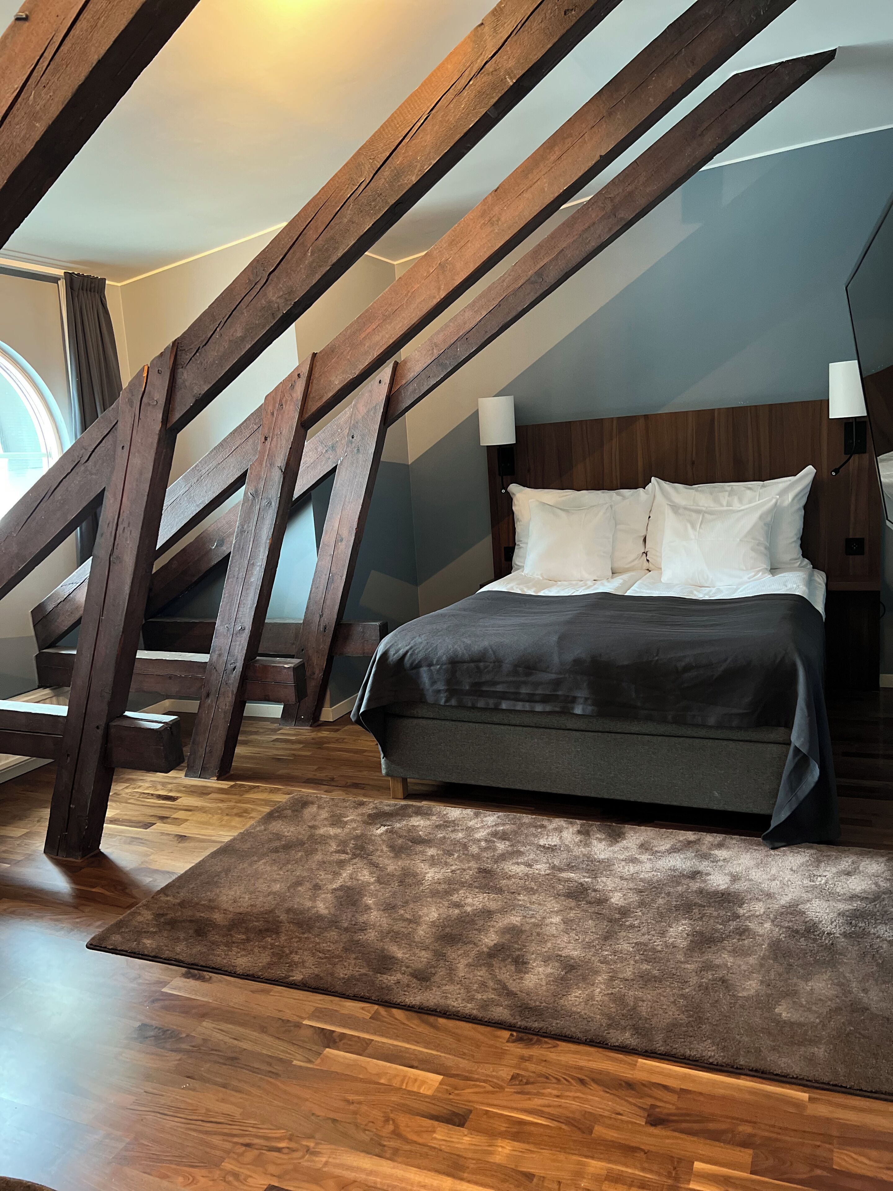 A bedroom with a sloped ceiling, wooden beams, a bed with white and dark bedding, and a window with curtains.