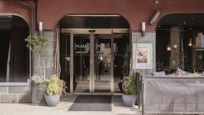 Hotellin sisäänkäynti, jossa on lasiovi, ruukkukasveja ja kyltti, jossa lukee: ”PLAZA HOME HOTEL”.