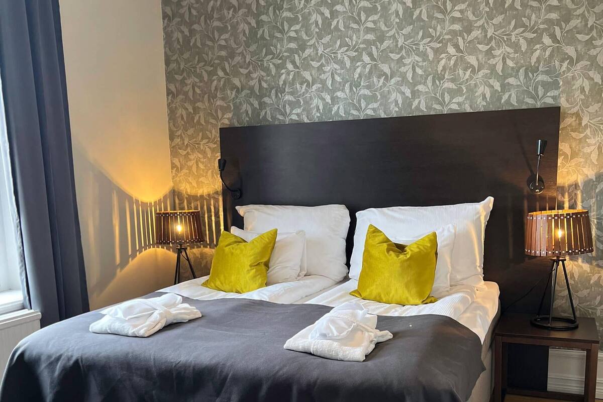 A neatly made bed with white and yellow pillows, a dark headboard, and a bedside table with a lamp.