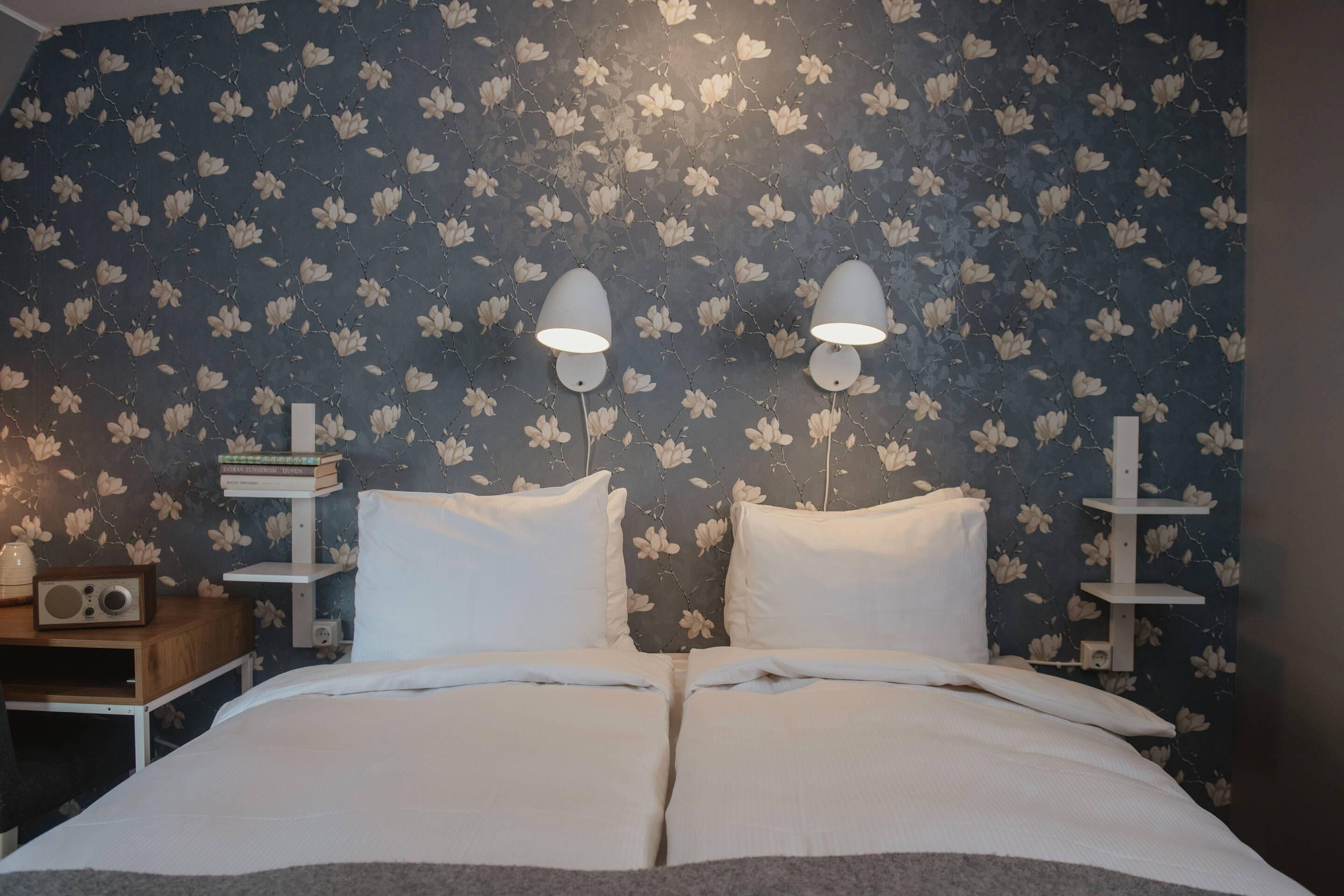 A neatly made bed with white bedding, two wall-mounted lamps, a bookshelf with books, and a floral wallpaper pattern.