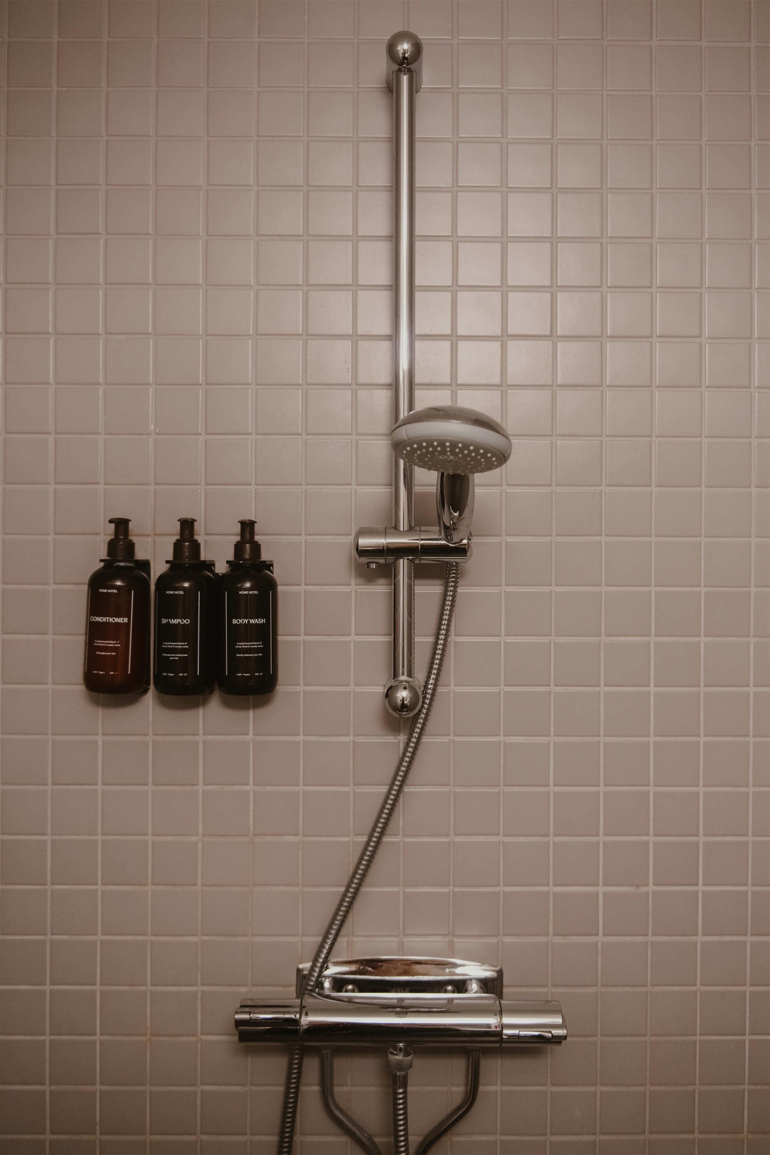A shower with a handheld showerhead and a wall-mounted control valve. Three bottles of toiletries are visible on the wall.