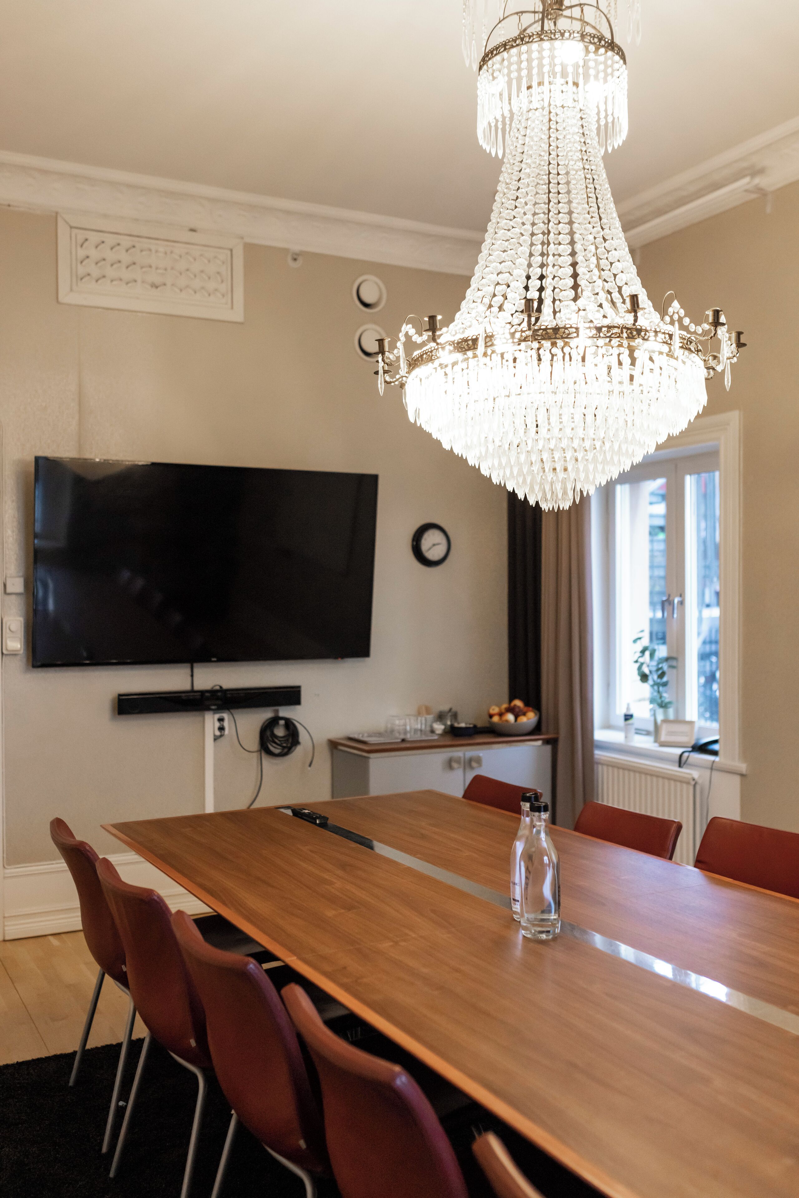 A dining area with a wooden table, red chairs, a chandelier, a flat-screen TV, and a window with a view.