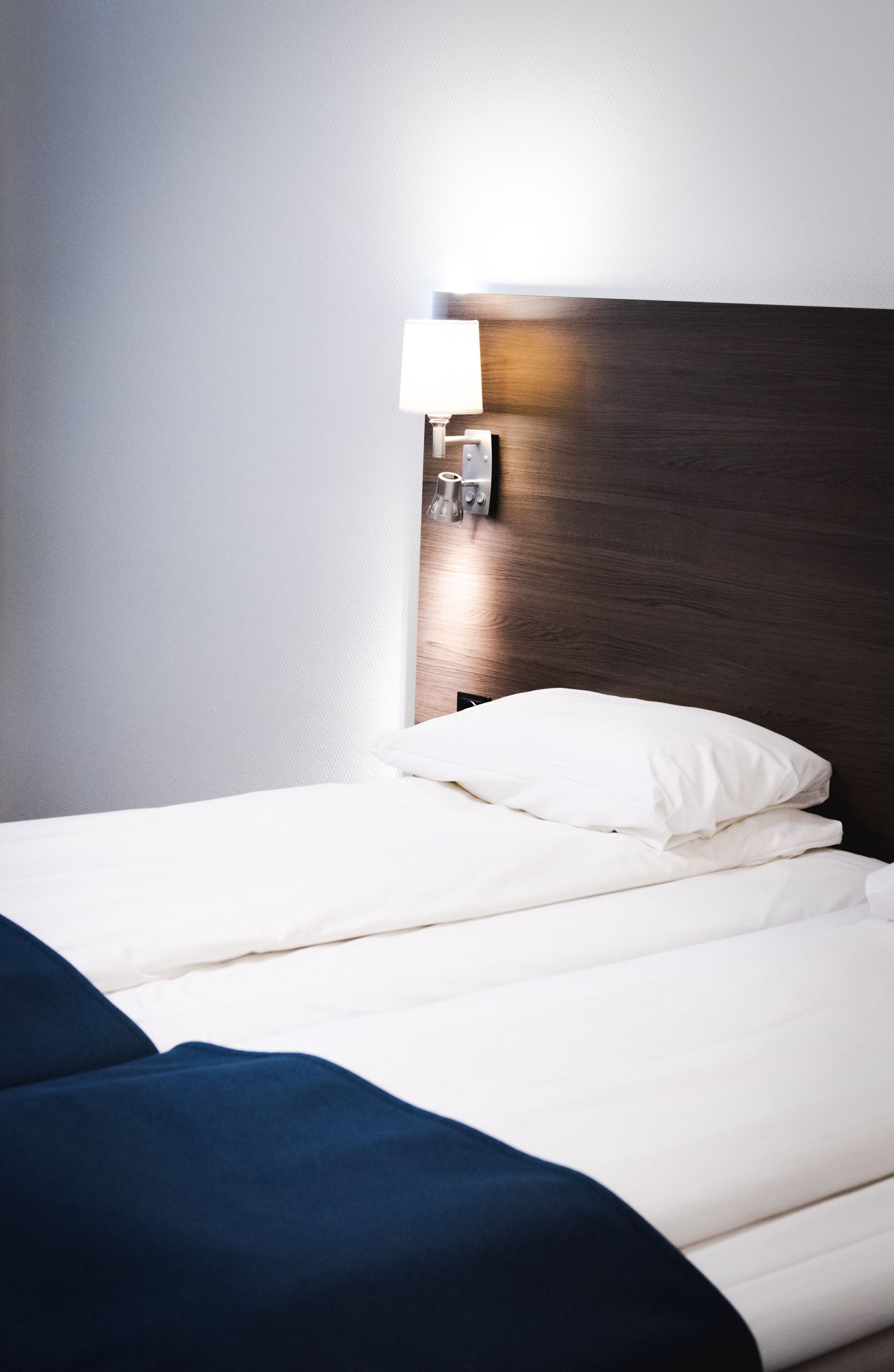 A neatly made bed with white bedding and a dark blue blanket, a wooden headboard, and a wall-mounted light fixture.