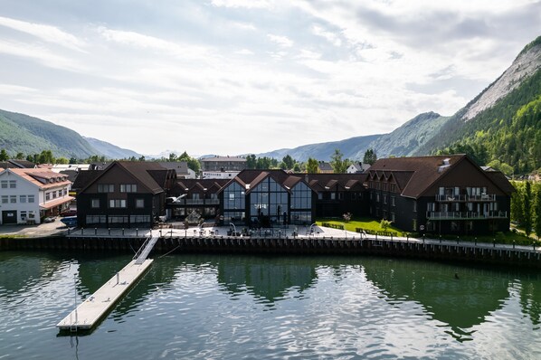 A lakeside hotel with a dock extending into the water, surrounded by mountains and greenery.