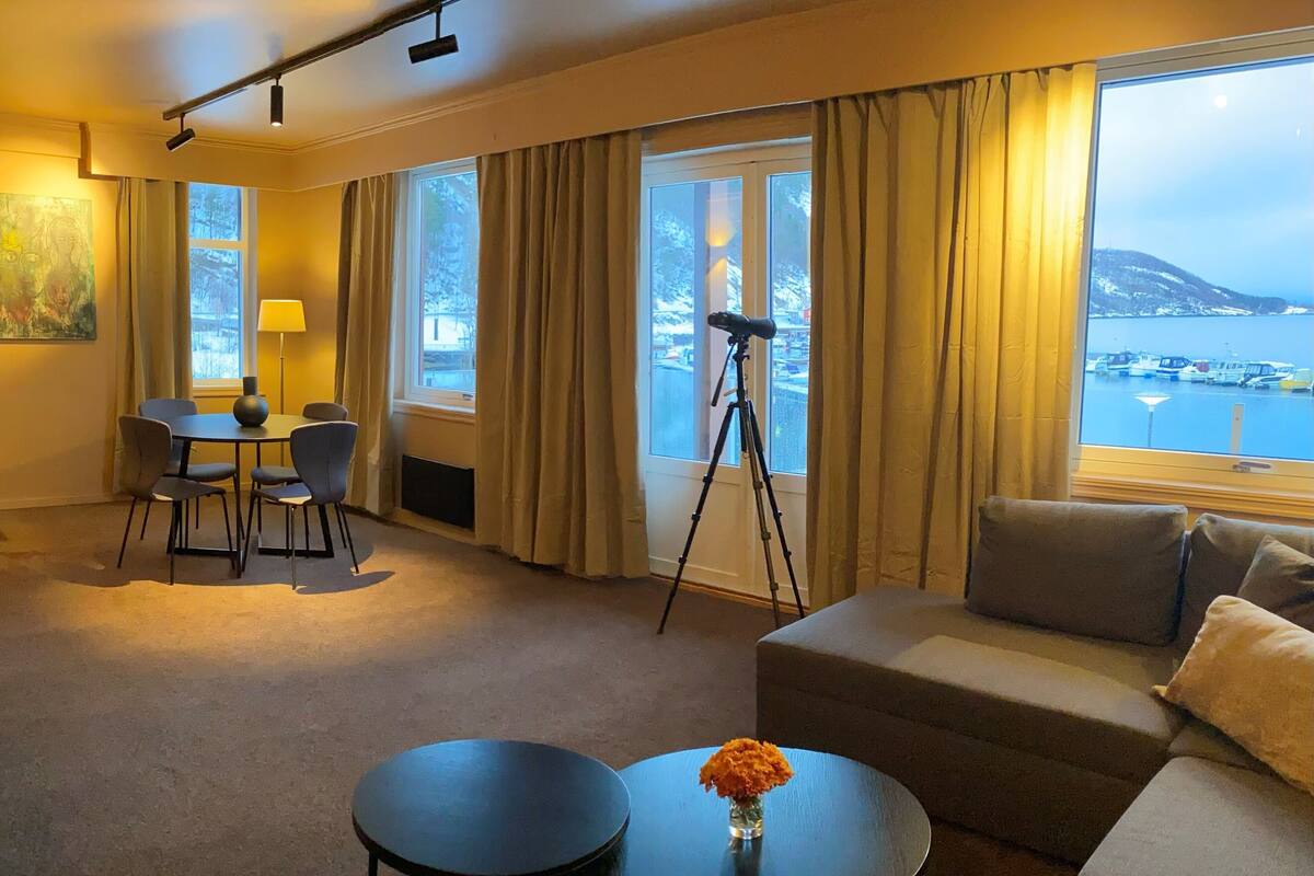 Una habitación de hotel moderna con sofá, dos mesas redondas, zona de comedor con sillas, telescopio y un amplio ventanal con vistas a barcos y un paisaje montañoso.