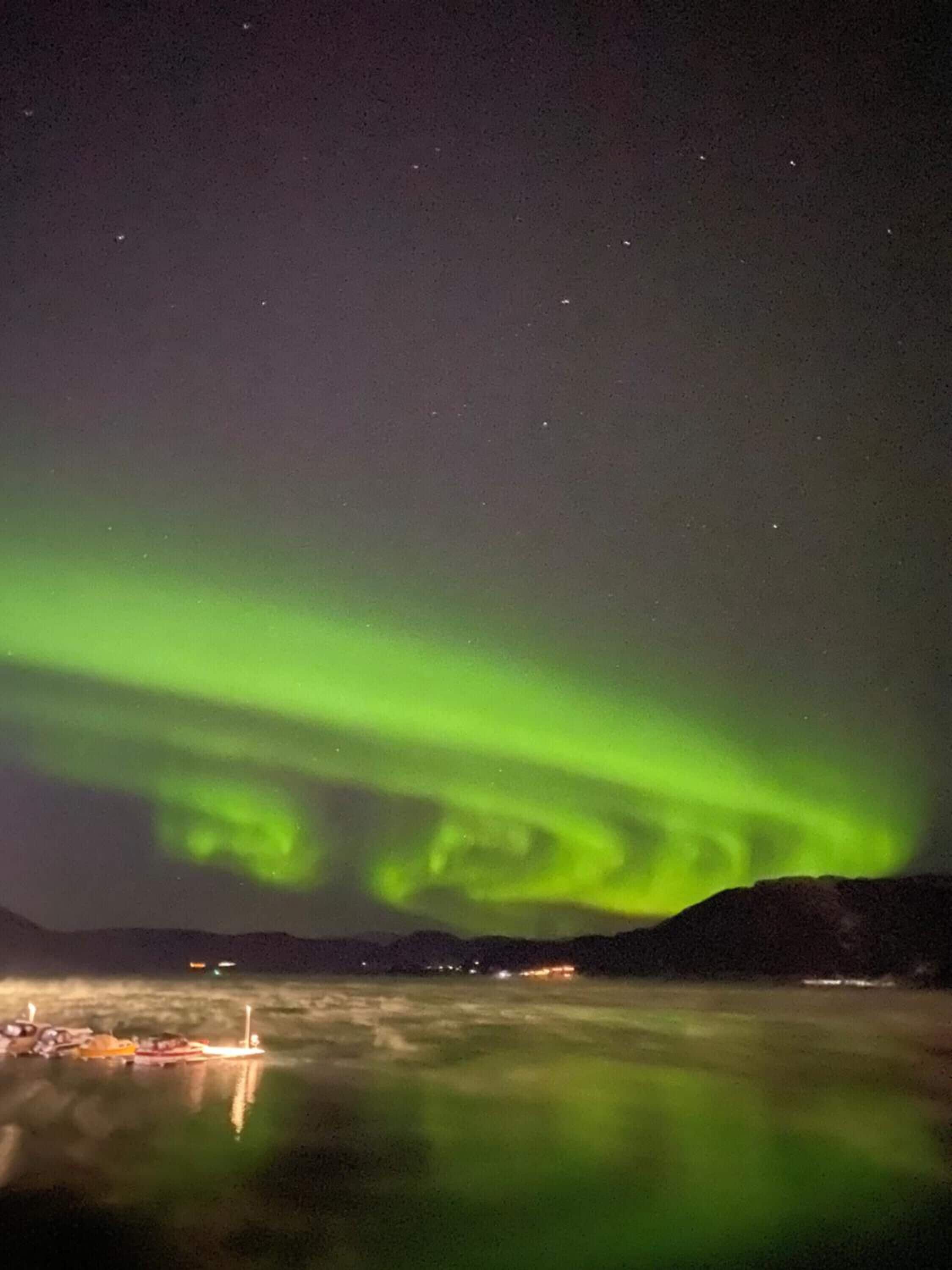 Vista nocturna de las auroras boreales sobre una masa de agua tranquila, con barcos y luces distantes.