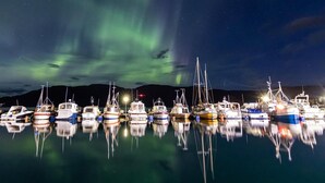 Un port de plaisance où les bateaux sont amarrés la nuit, se reflétant sur l’eau calme, sous un ciel étoilé où l’on peut apercevoir des aurores boréales.