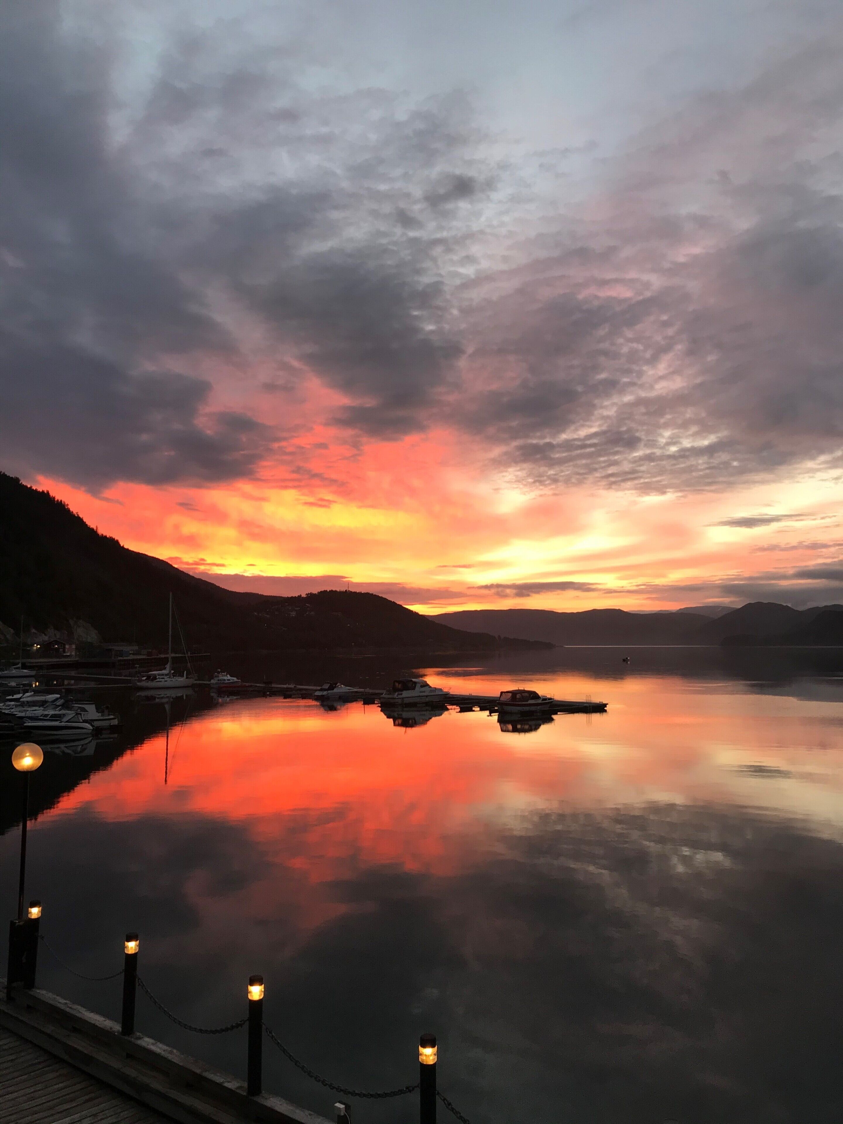 Un lago tranquilo al atardecer con barcos amarrados, un muelle iluminado y un paisaje montañoso al fondo.