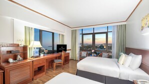 Een hotelkamer met een groot bed, een bureau met een computer, een stoel en uitzicht op de stad door grote ramen.