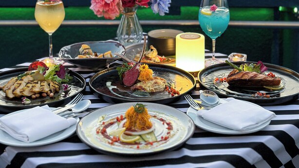 Mesa dispuesta con varios platos, bebidas, un jarrón de flores y una vela, con vista a un paisaje urbano al atardecer.