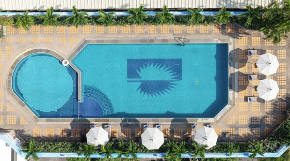 Vista aérea de una piscina rectangular con un diseño de sol, rodeada de palmeras y sombrillas.