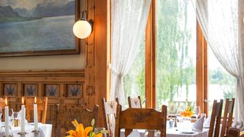 A dining table set with candles, flowers, and glassware, with a view of greenery and a painting on the wall.