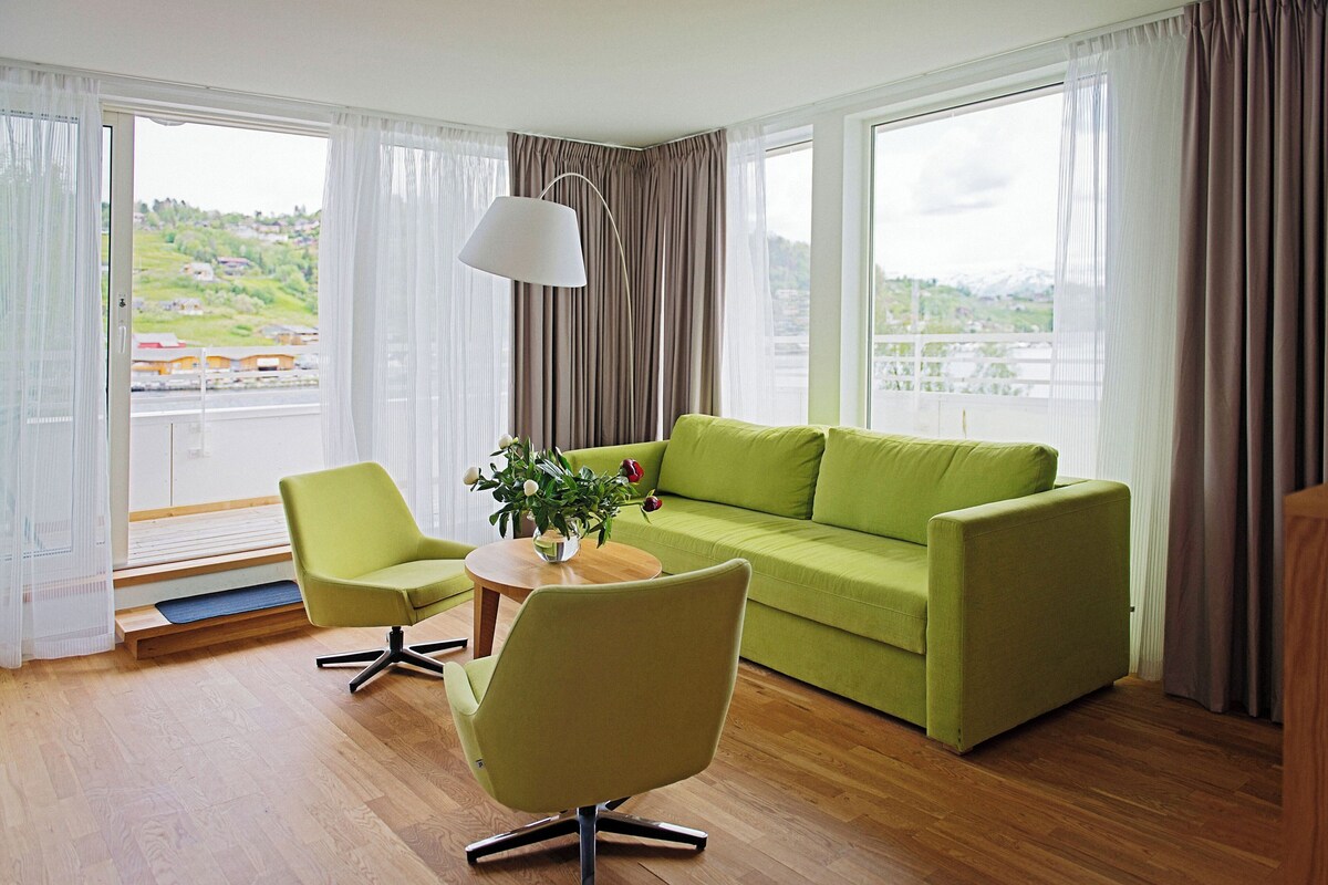 A modern living room with a green sofa, two armchairs, a wooden coffee table, and a view of a cityscape through large windows.