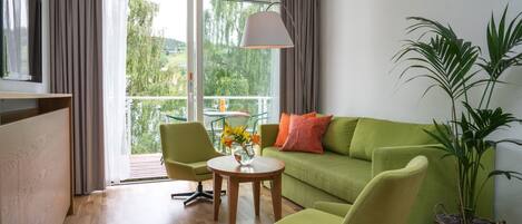 Una sala moderna con un sofá verde, dos sillones a juego, una pequeña mesa de madera y una lámpara de pie. Un ventanal grande ofrece vistas al verdor exterior.
