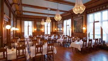 A spacious dining area with multiple tables and chairs, wooden floors, and large windows with paintings.