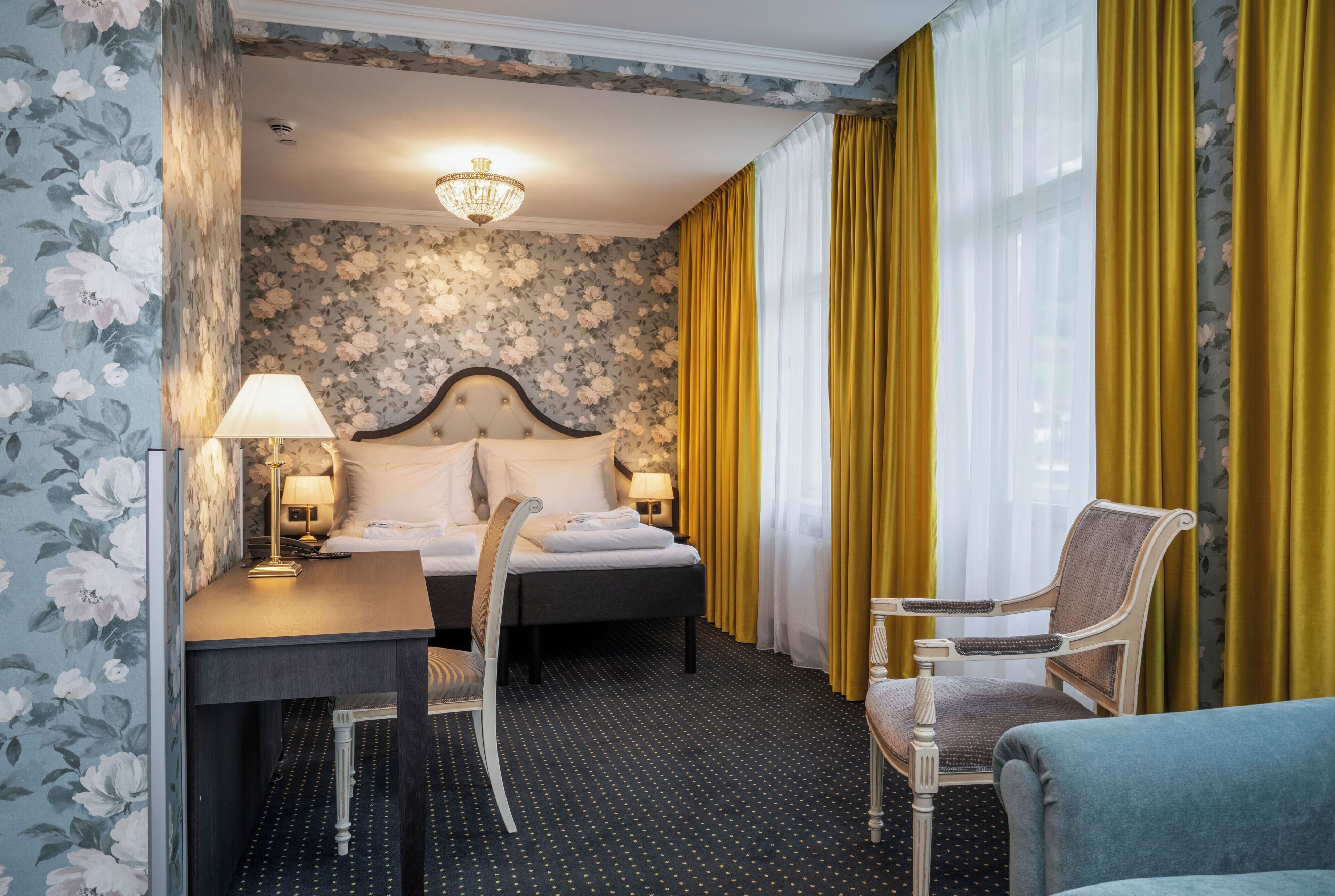 Hotellihuone, jossa on sänky, kirjoituspöytä lampun kanssa, tuoli ja sohva.