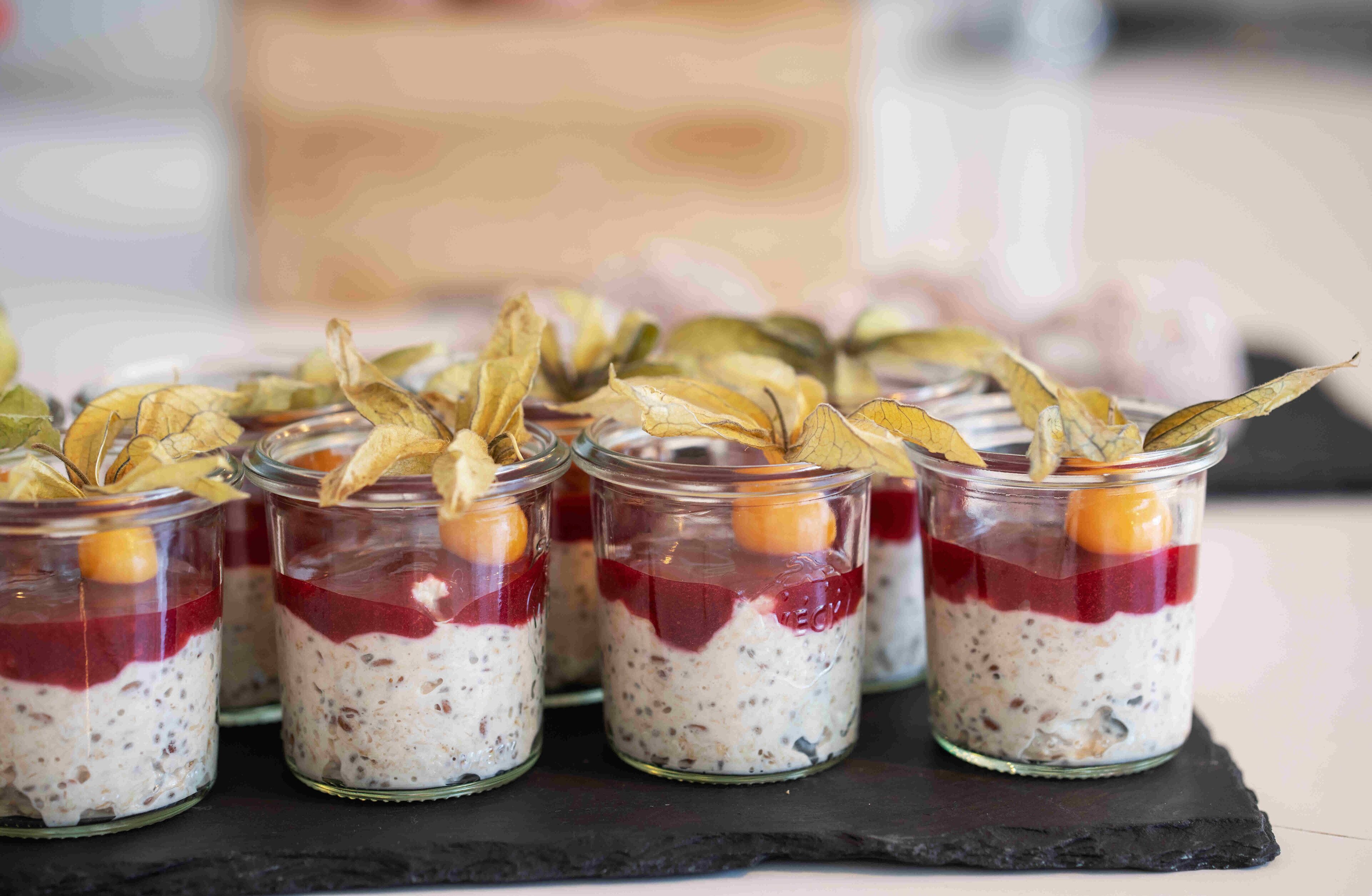 Une série de bocaux en verre contenant des aliments disposés en couches, chaque bocal étant surmonté d’une feuille séchée et d’un petit fruit.