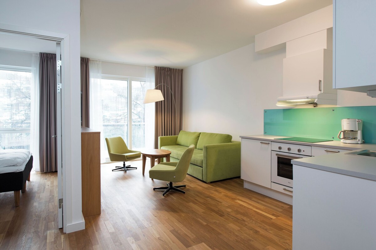 A modern hotel room with a green sofa, two armchairs, a small table, a kitchenette, and a bed.