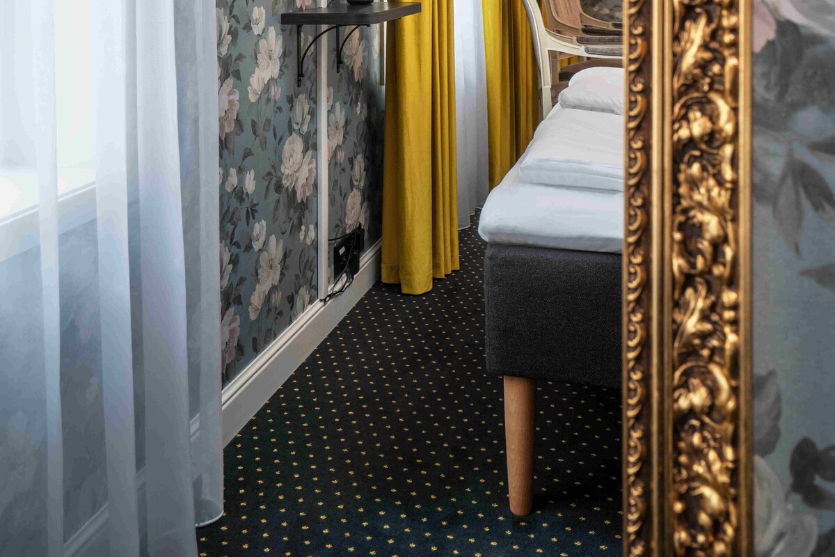 Habitación de hotel con papel tapiz floral, una cama, una mesita de noche con lámpara, un espejo y un marco de espejo grande y ornamentado.