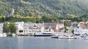 A marina with boats docked, surrounded by traditional buildings and a forested hill in the background. - Thon Hotel Sandven (Kvam)