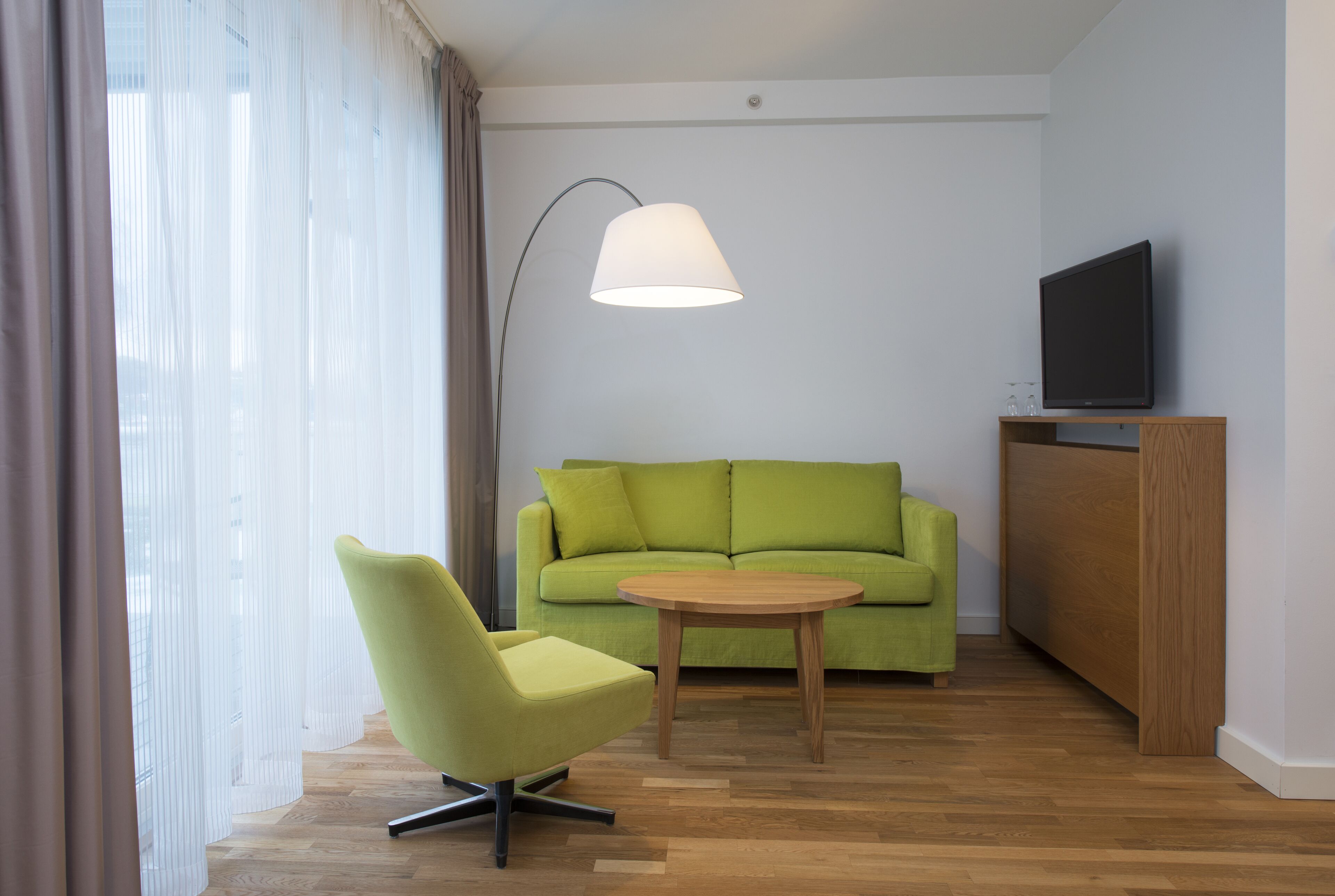 A modern room with a green sofa, a wooden coffee table, a green armchair, a TV on a wooden stand, and sheer curtains on the window.