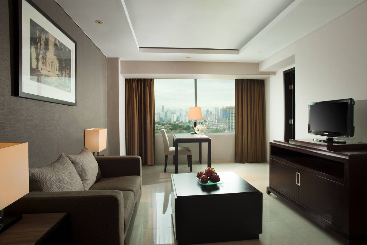 A modern hotel room with a sofa, a coffee table, a TV, and a city view.