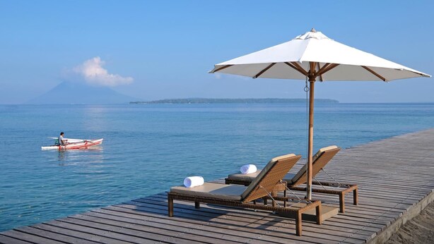 A wooden deck with lounge chairs and a white umbrella, overlooking a calm sea with a boat and distant land. - Grand Luley Manado (Manado)