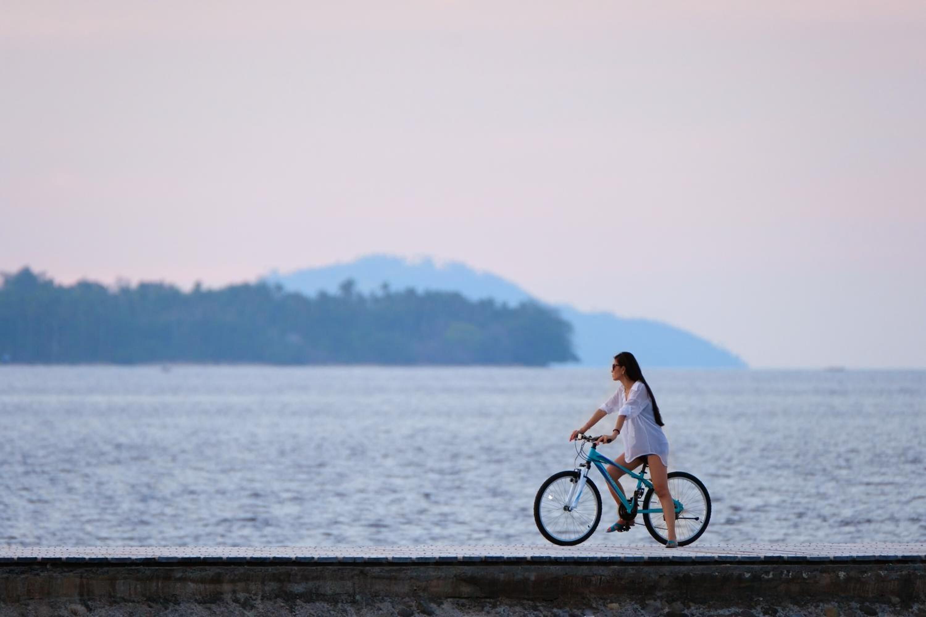 Una persona pedaleando en bicicleta por un camino de concreto junto a una masa de agua, con tierra a lo lejos.
