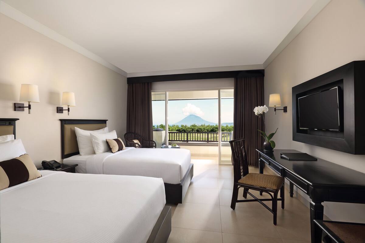Ein Hotelzimmer mit zwei Betten, einem Schreibtisch, einem Stuhl, einem Fernseher und einem Balkon mit Bergblick.