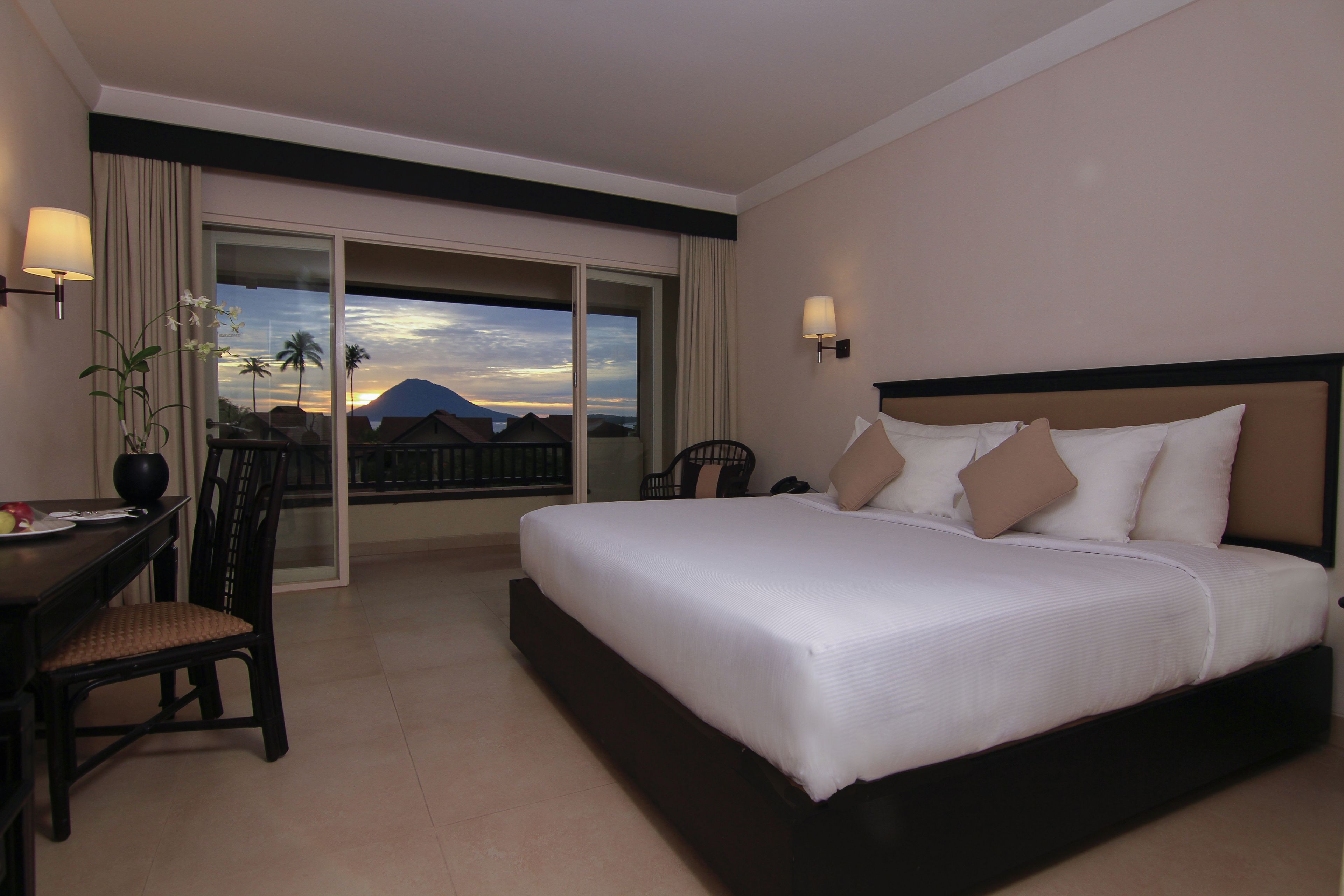 Habitación de hotel con una cama grande, un escritorio con una silla, una planta en maceta y vistas a un atardecer sobre una montaña.