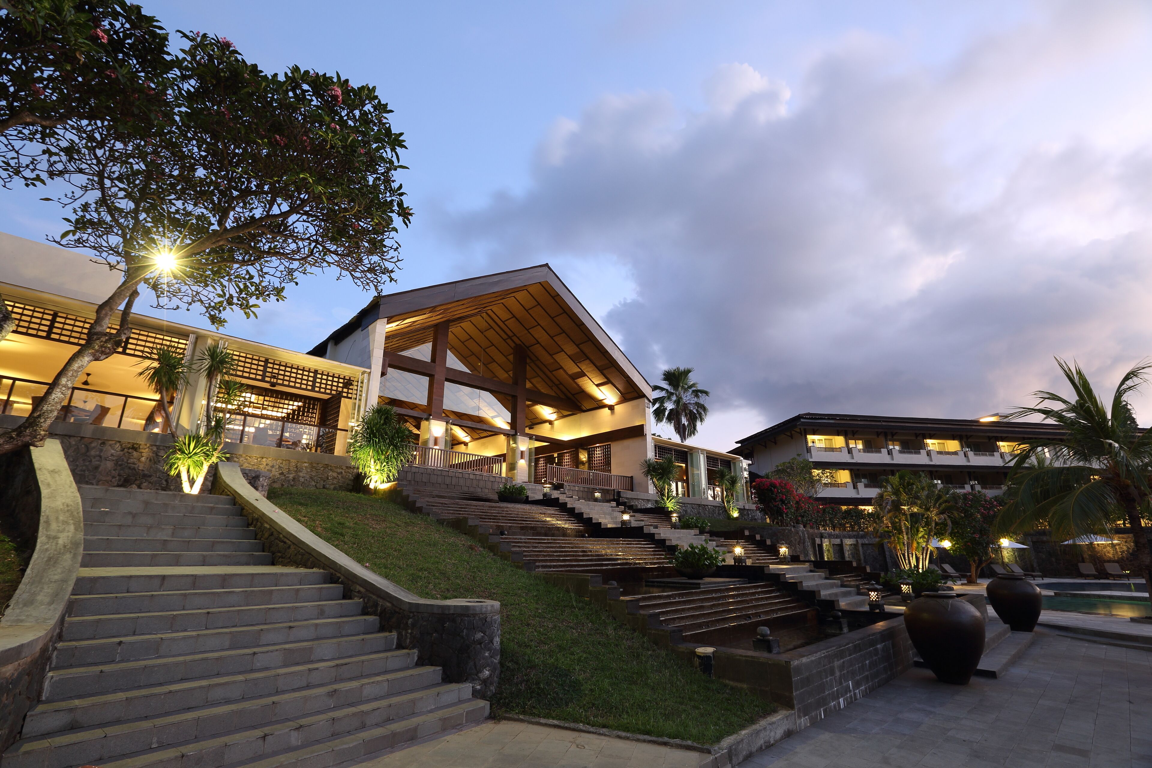 A hotel with a modern architectural design, featuring a prominent staircase leading to the entrance, a pool area, and lush landscaping.