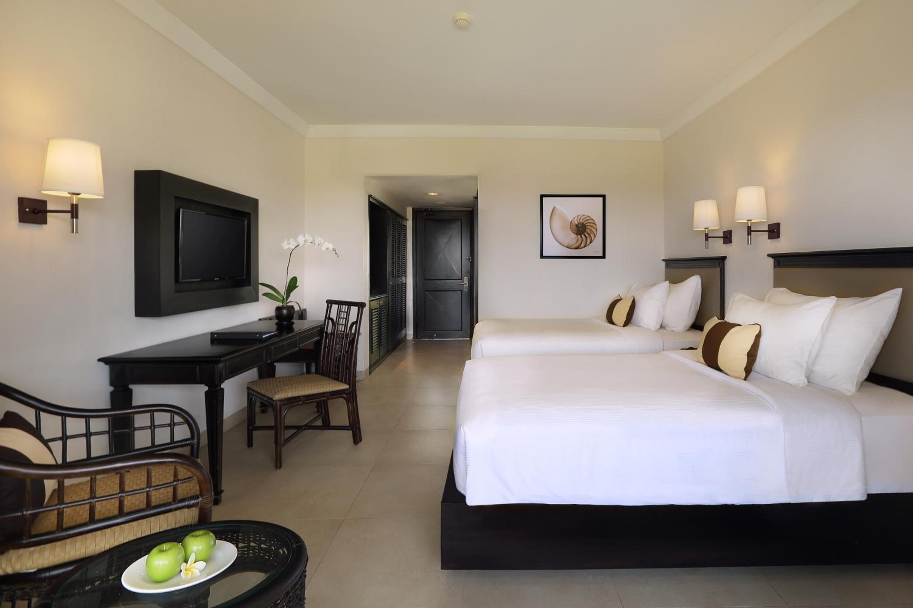 Ein Hotelzimmer mit zwei Betten, einem Schreibtisch, einem Stuhl, einem Fernseher und einem Esstisch mit Äpfeln und einer Blume.