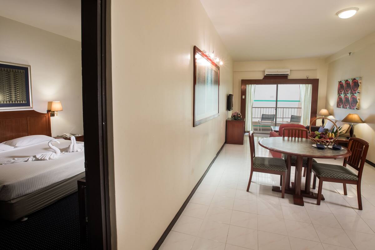 A hotel room with a bed, a dining table with chairs, a small table with a lamp, and a large mirror.