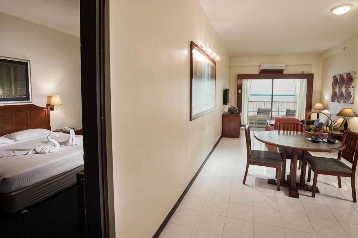 A hotel room with a bed, a dining area with a table and chairs, a view of the balcony, and a wall-mounted TV.