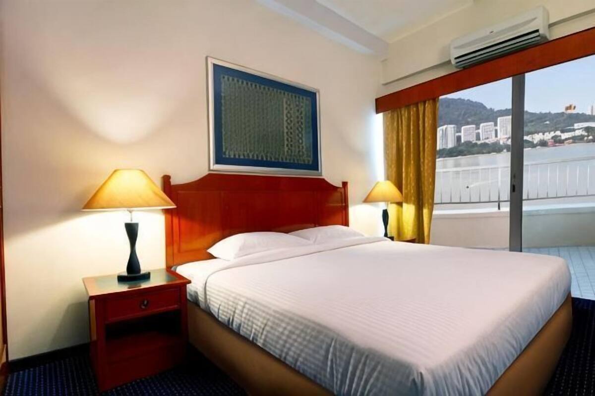 A hotel room with a bed, two bedside lamps, a nightstand, a window with curtains, and a view of buildings.