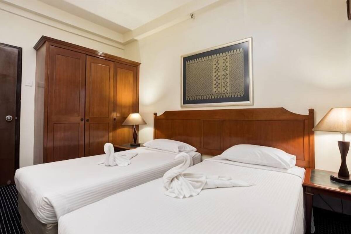 A hotel room with two beds, a wooden wardrobe, a decorative wall hanging, and bedside lamps.